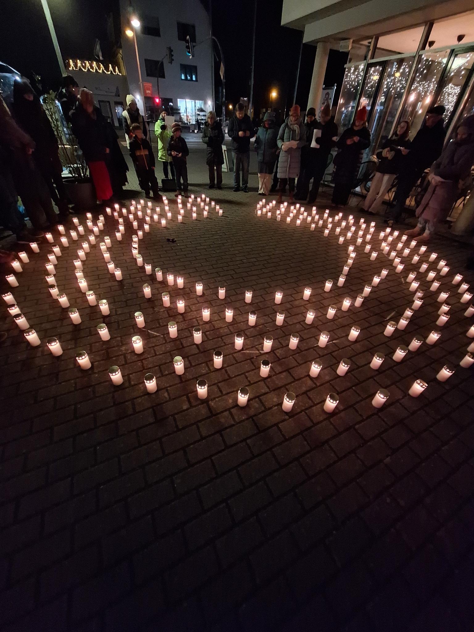 Vier Kreise mit brennenden weißen Grablichtern. Darum herum sieht man einige Menschen